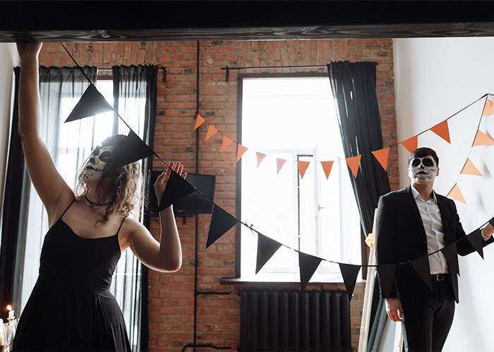 Woman and man in skull Halloween costumes hanging black and orange decorations for an inappropriate costume party.