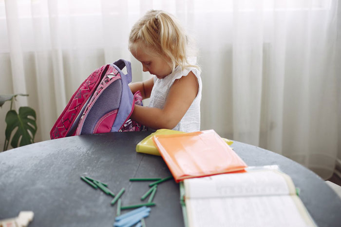 Young girl packing a backpack at a table with school supplies, reflecting a single mom&rsquo;s plastic surgery support dilemma.