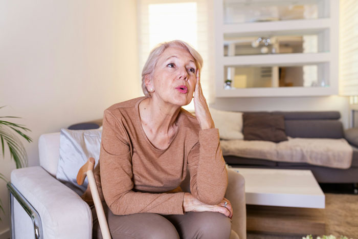 Older woman sitting on chair at home, looking thoughtful and stressed, relating to single mom plastic surgery support struggles.