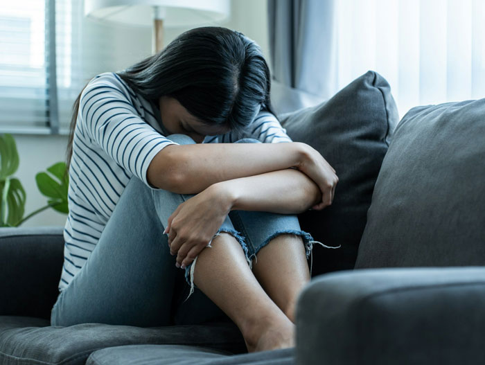 Single mom sitting on couch looking distressed after spending thousands on plastic surgery with no support.