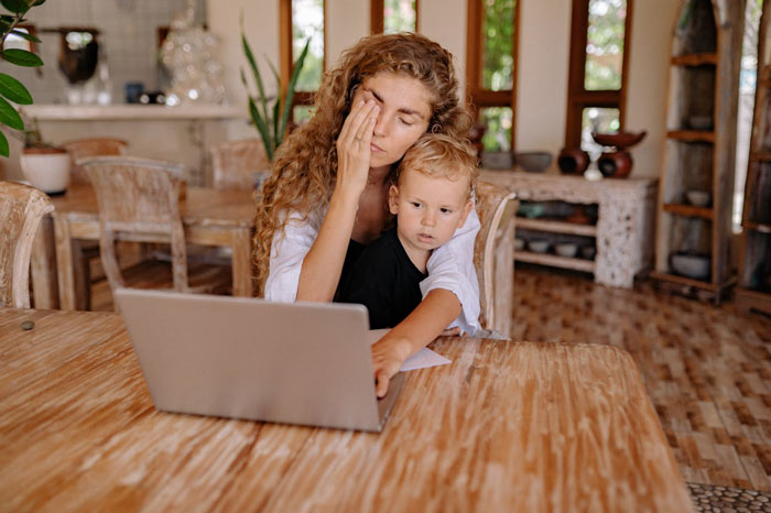 Stressed mother working on laptop with child on lap, reflecting challenges of parents guilt-tripping and cheap babysitting refusal.