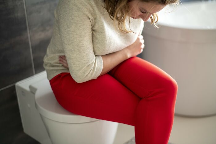 Woman in red pants sitting on a toilet, holding her stomach in pain, illustrating severe medical problems and discomfort.