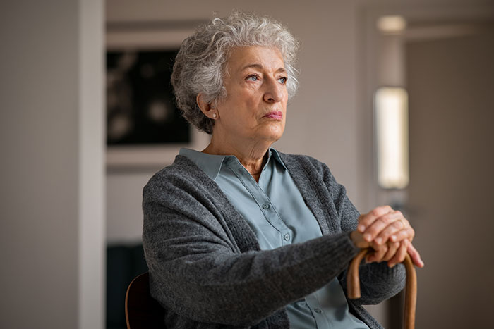 Elderly woman with gray hair sitting indoors, looking serious and contemplative about financial ruin issues.