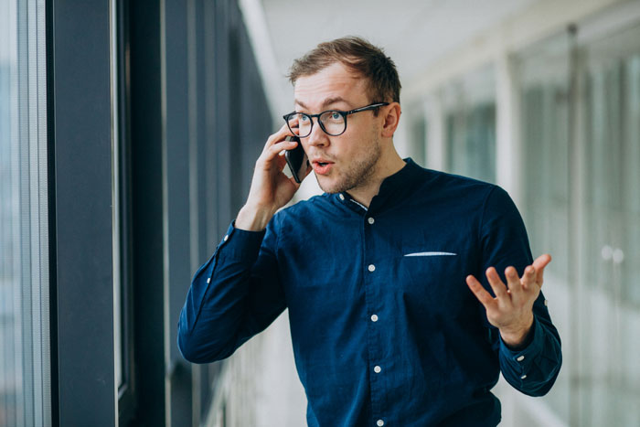 Man in glasses and blue shirt talking on phone in office hallway, showing quiet gossip drama in workplace.