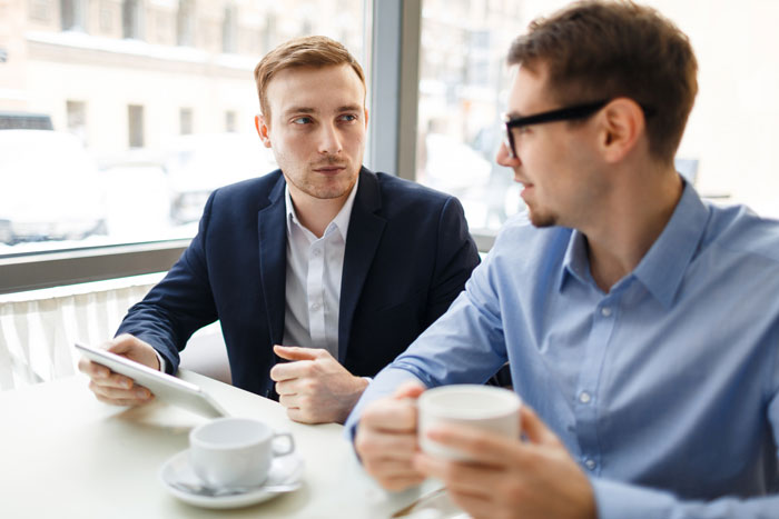 Two young men having a quiet work conversation over coffee, possibly discussing gossip or drama during a break.
