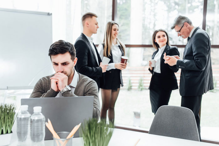 Man working quietly on laptop in office while coworkers engage in gossip and drama behind him.