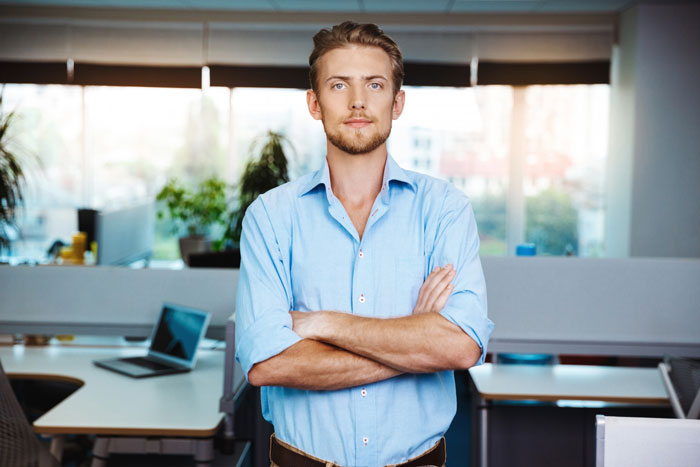 Young man standing with arms crossed in a modern office, embodying work quiet focus away from gossip and drama.