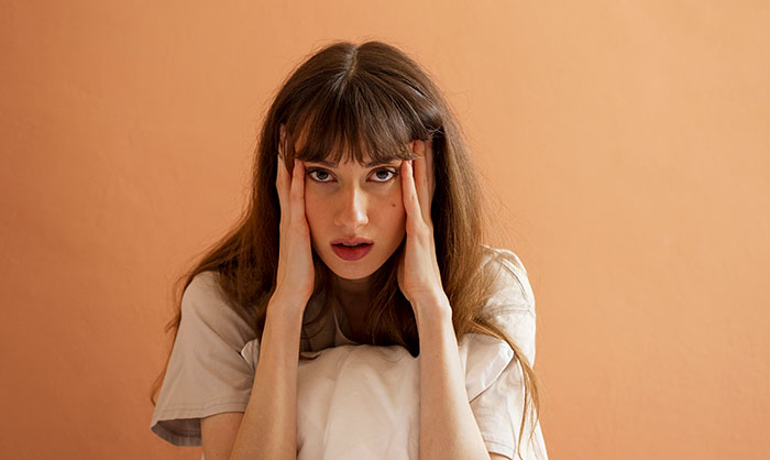 Young woman babysitter looking stressed and overwhelmed while sitting against a peach wall, reflecting awkward moments.