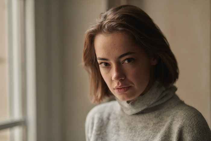 Young woman babysitter in a gray turtleneck, looking thoughtful and reflective by a window indoors.
