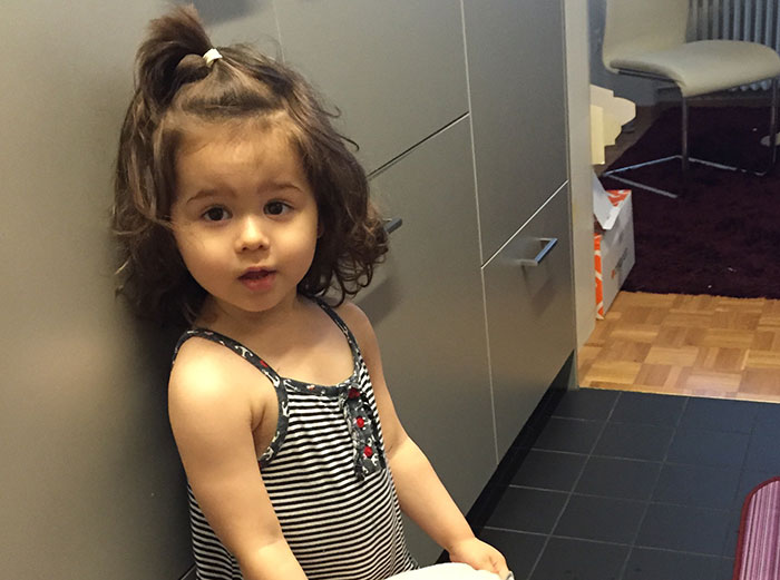 Young toddler girl with curly hair wearing a striped dress standing in a kitchen, relating to babysitters weird moments.