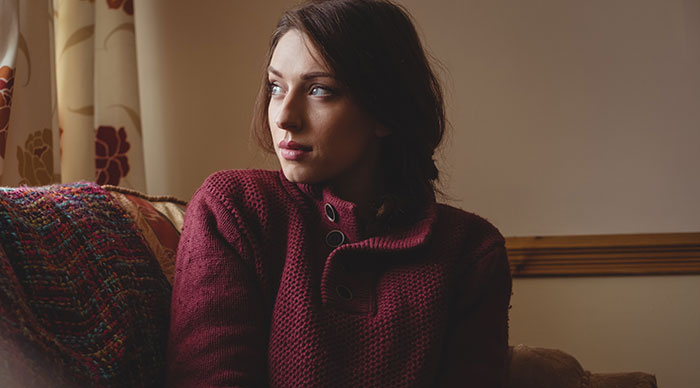 Young babysitter in a red sweater sitting on a couch, looking thoughtfully out the window during a quiet moment.