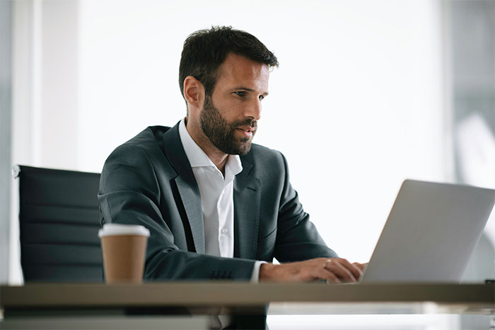 Man in a business suit working on a laptop at office desk with coffee cup, illustrating times people failed spectacularly at work.