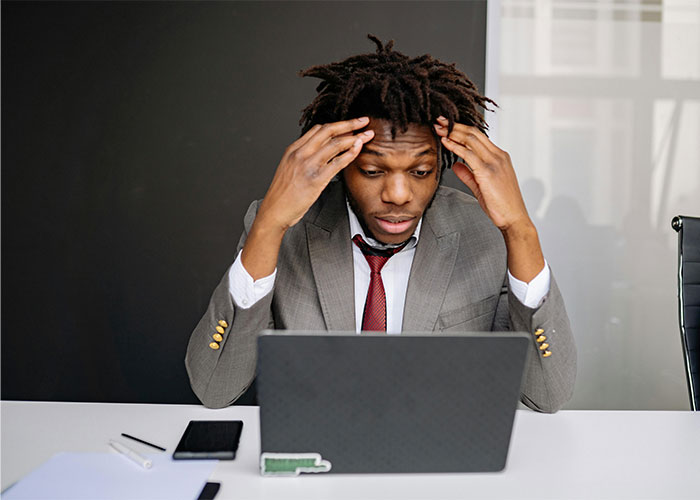 Stressed man in a gray suit sitting at a desk looking at laptop, illustrating people who failed spectacularly at work.