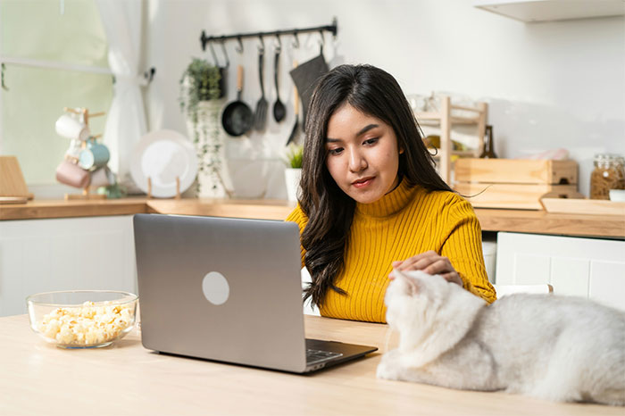 Woman working on laptop while petting white cat at kitchen table, showcasing relatable work moment for people failed spectacularly at work.
