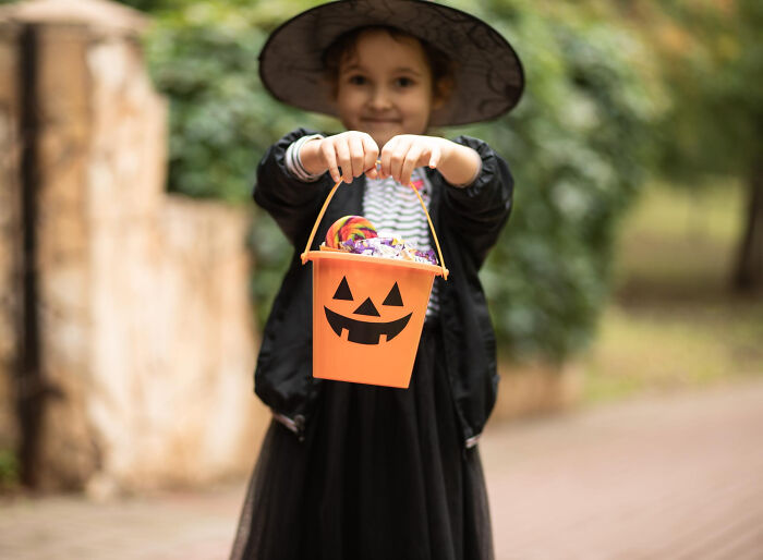 Young girl in a witch costume holding a pumpkin bucket filled with Halloween candy, capturing a spooky moment.