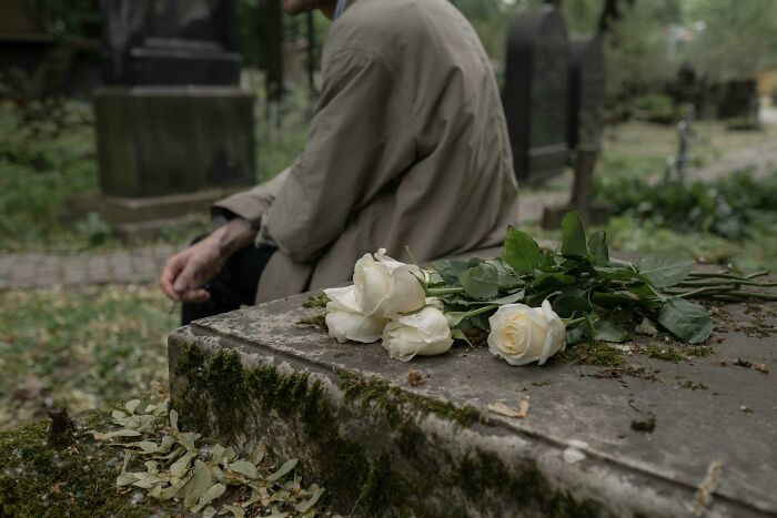 Person in a graveyard sitting beside a tombstone with white roses, illustrating mind-bending scenarios that challenge your morals.