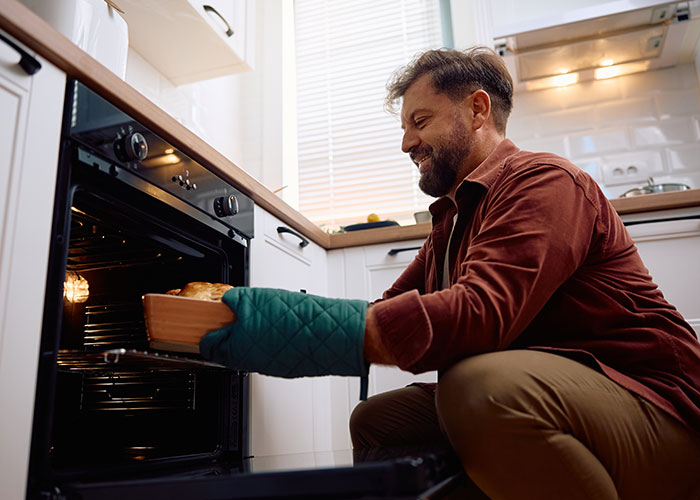 Man wearing oven mitts, smiling while taking out freshly baked bread, illustrating myths about men cooking at home.