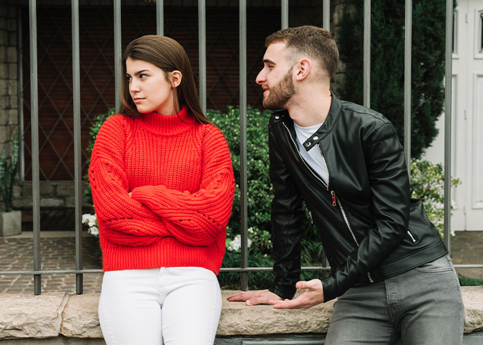 A man in a black jacket shares myths about men while a woman in a red sweater listens with crossed arms outside by a fence.