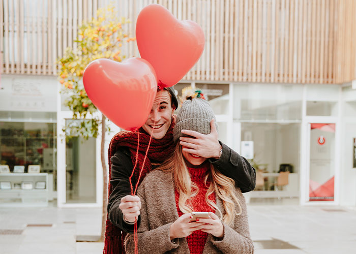 Young man holding heart-shaped balloons and covering a woman’s eyes, symbolizing men sharing myths about them that are not true.