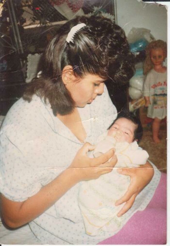 Woman feeding a baby with a bottle while a creepy doll with glowing eyes stands in the background, eerie photo moment.