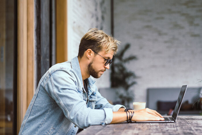 Young man wearing sunglasses and denim jacket working on a laptop, focusing on finding loopholes to take advantage of now.