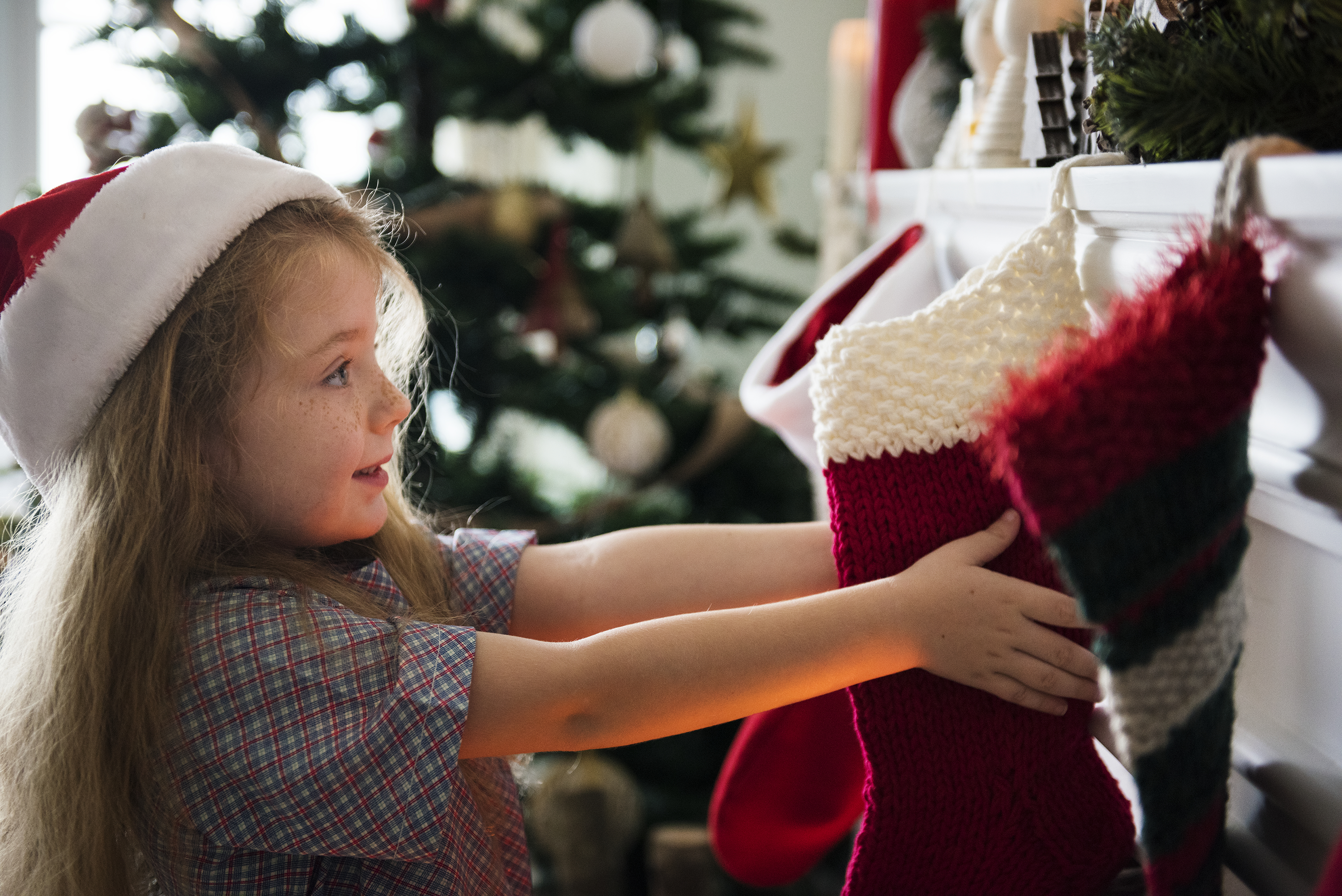 Young girl in Santa hat hanging Christmas stockings by the fireplace during holiday decorating with festive tree in background
