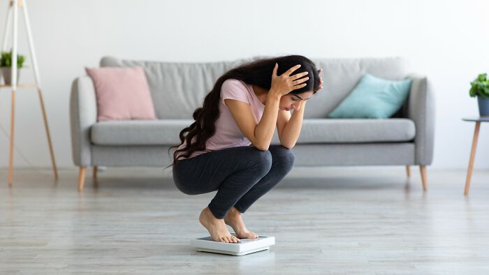 Woman stressed about medical problems, crouching on a scale in a living room with a gray couch and cushions.