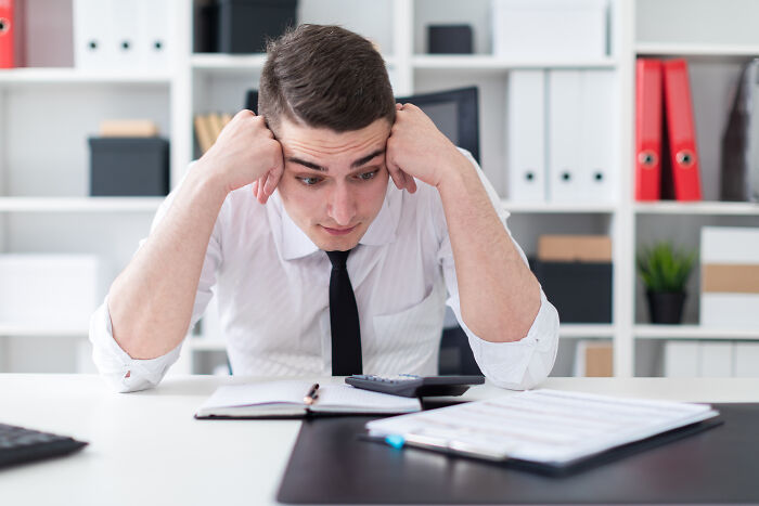 Young accountant in white shirt and black tie looking stressed while working with calculator and documents in office setting