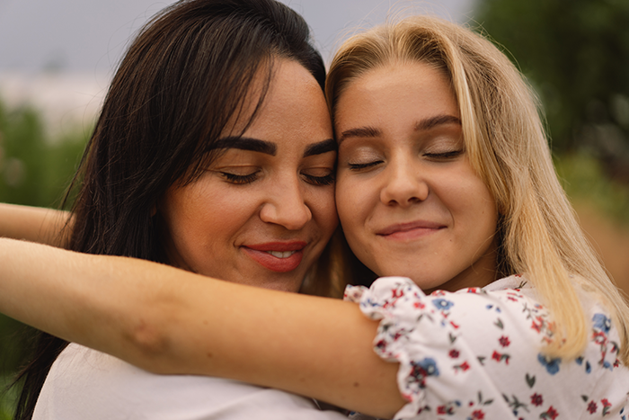 Close-up of a happy clean-f***k mom and daughter sharing an embrace outdoors with eyes closed and content smiles.