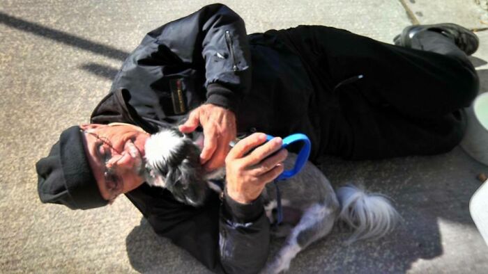 Man lying on the ground, smiling and playing affectionately with a small dog, capturing faith in humanity restored moment.