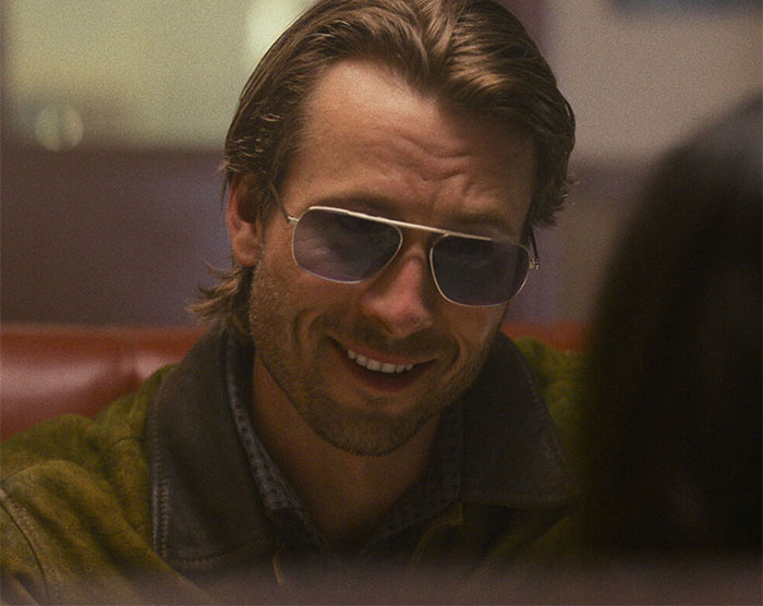Glen Powell wearing sunglasses and smiling during an interview, highlighting viral blunder and dark side of Hollywood.