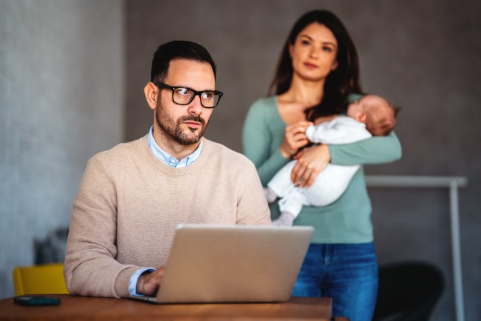 Man prioritizes work at laptop while wife stands behind holding baby, showing emotional distance in family. Man prioritizes work at laptop while wife stands behind holding baby, showing emotional distance in family.
