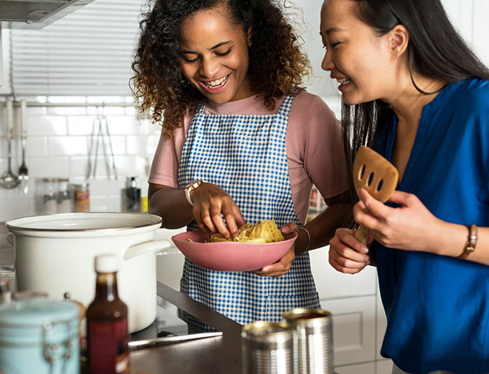 Two women cooking wild American food creations in a kitchen, preparing a dish with noodles and smiling together. Two women cooking wild American food creations in a kitchen, preparing a dish with noodles and smiling together.