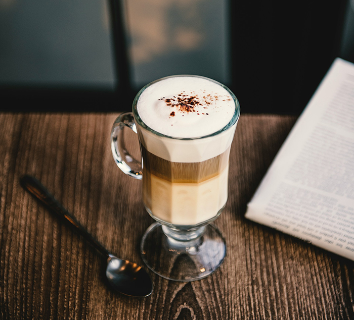 Glass of morning coffee with foam and chocolate sprinkles on a wooden table next to a newspaper and spoon
