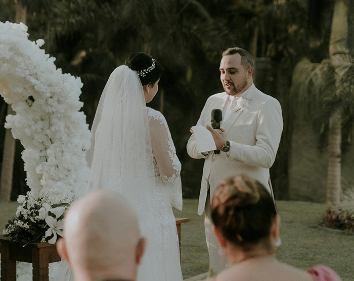 Groom reading wedding vows with microphone at outdoor ceremony, bride standing nearby in lace dress and veil.