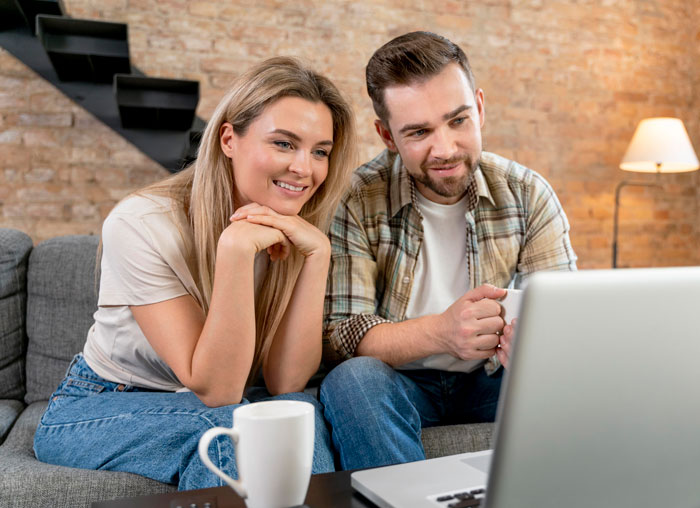 Couple looking at laptop together on couch, discussing vacation plans while dealing with MIL joining their trip.