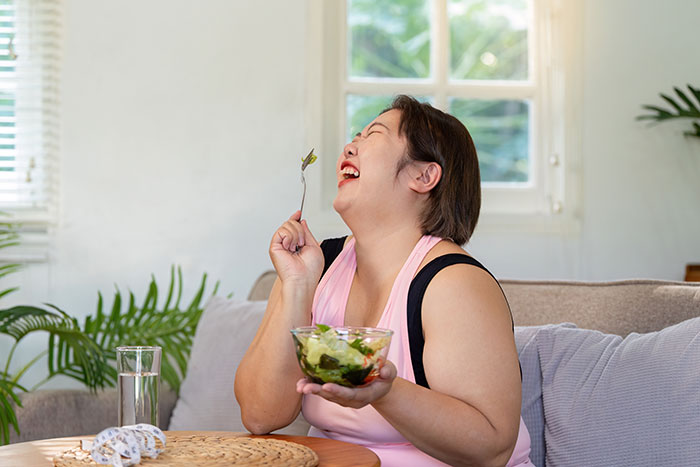 Happy woman eating salad at home after gaining at least 40 lbs, sitting on a couch near a window.