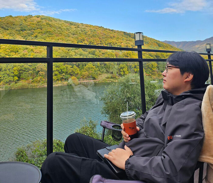 A man from South Korea wearing glasses and a jacket, sitting outdoors by a lake with autumn foliage.