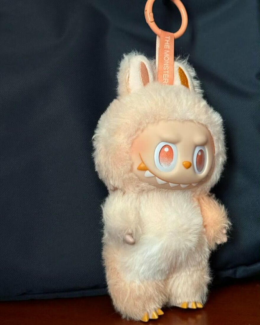 Original Labubu doll with fluffy fur and orange eyes hanging against dark fabric background, a hot property collectible.