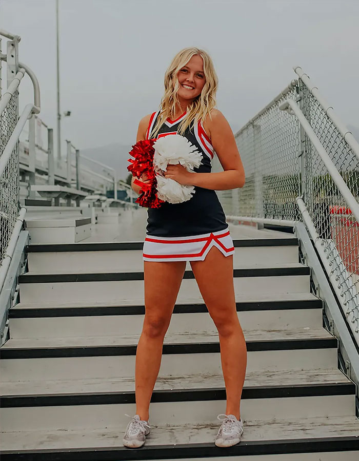 Teen girl who was paralyzed at 16 posing confidently in cheerleading uniform on stadium stairs holding pom-poms.