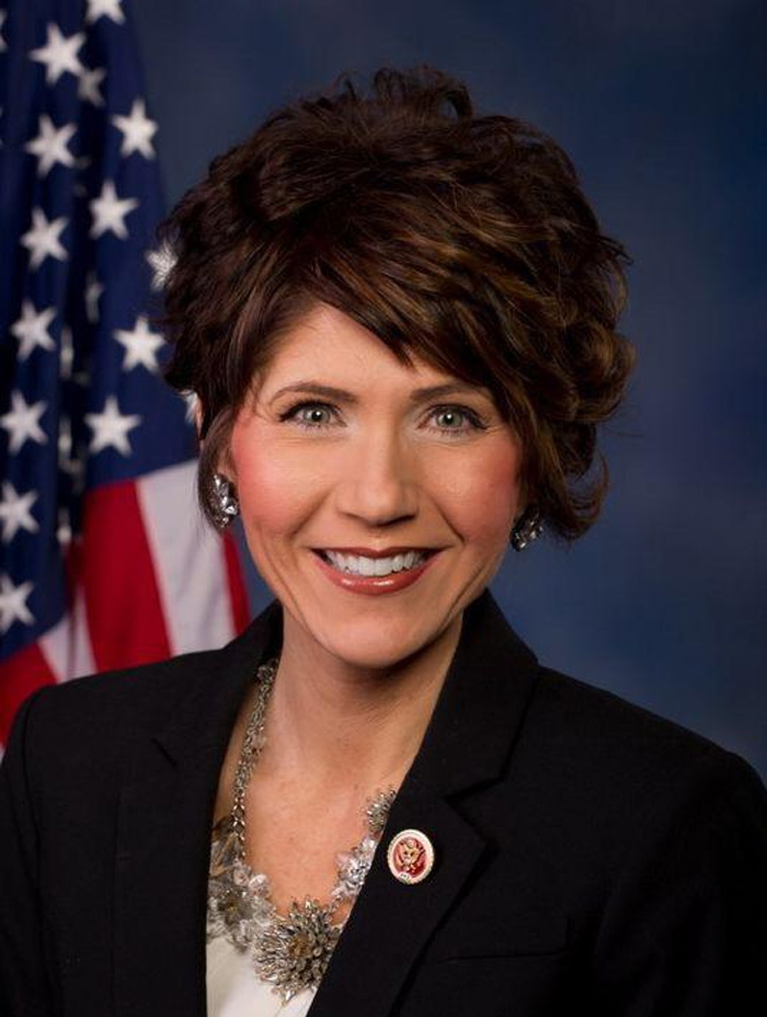 Kristi Noem smiling in a formal portrait with short styled hair and American flag in the background. Kristi Noem smiling in a formal portrait with short styled hair and American flag in the background.