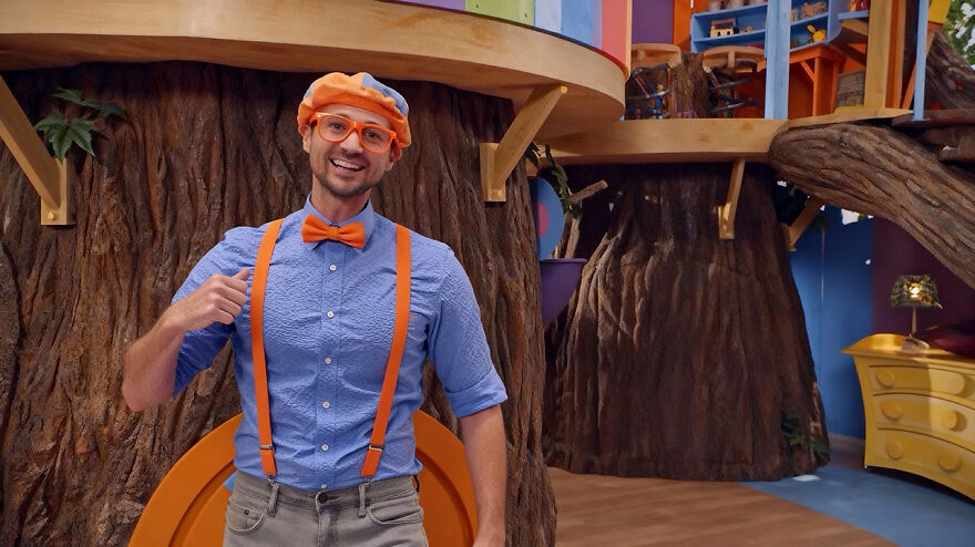 A Blippi actor smiling indoors wearing a blue shirt, orange bow tie, suspenders, and hat in a colorful set background.