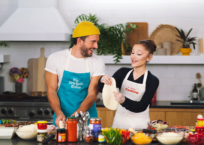 Ariana Grande and Jonathan Bailey sharing a lighthearted moment while preparing food in a modern kitchen.