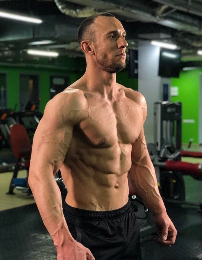 Muscular fitness coach posing confidently in gym, highlighting toned physique and vascular definition during workout session.