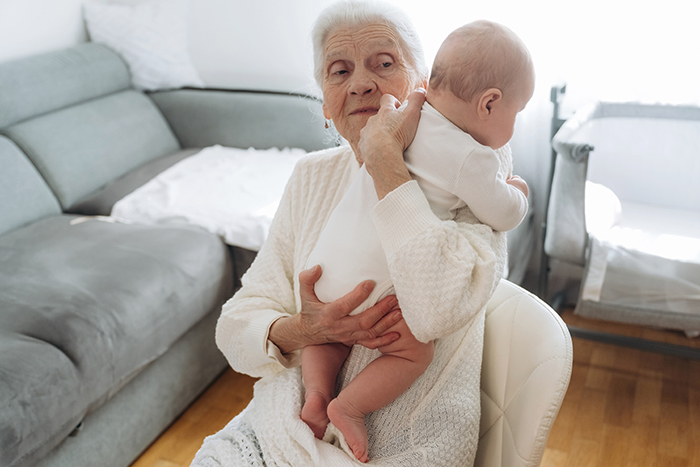 Elderly woman holding a baby in a living room, highlighting mom's concerns about leaving baby with mom or MIL alone.