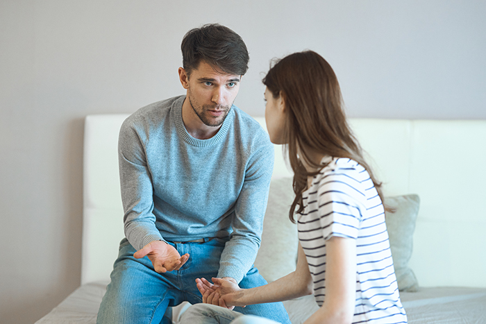 Husband bewildered talking to wife on bed, confused expression about wedding and marriage conversation.