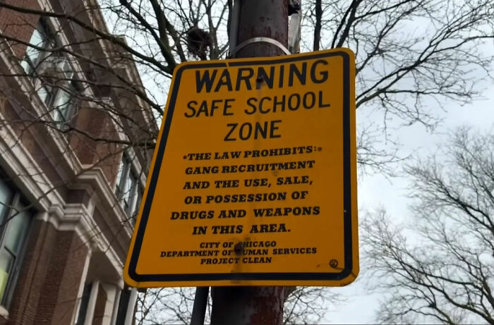 Yellow warning sign for safe school zone prohibiting gang recruitment, d***s, and weapons in an urban area near a building.