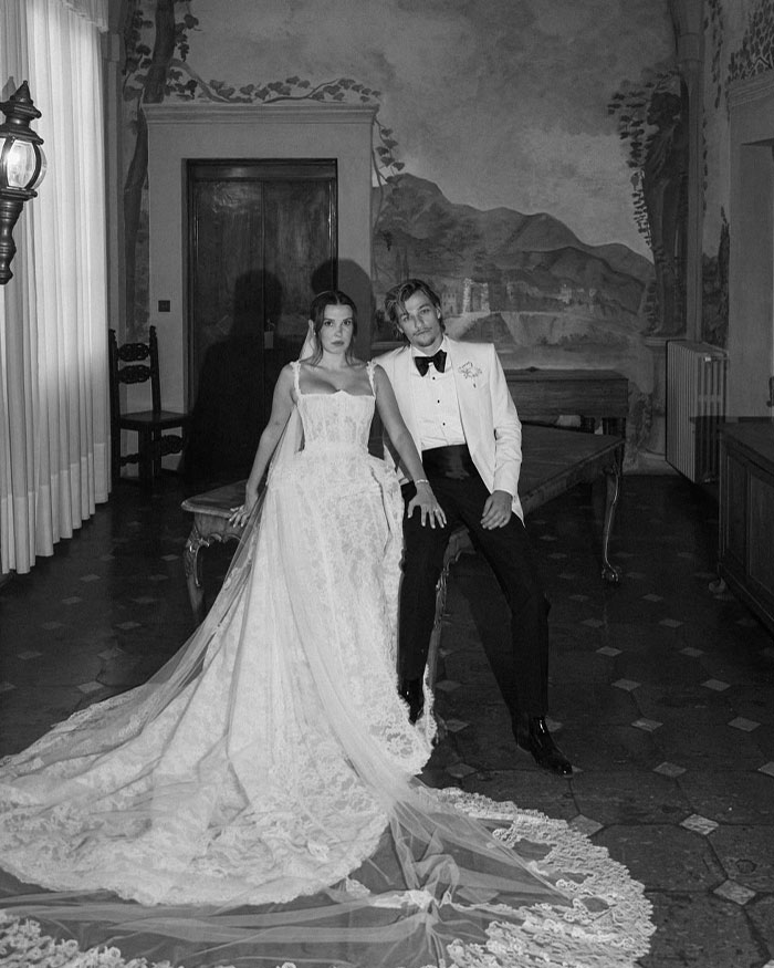 Black and white photo of a couple in formal attire, with Millie Bobby Brown wearing a long lace wedding dress indoors.