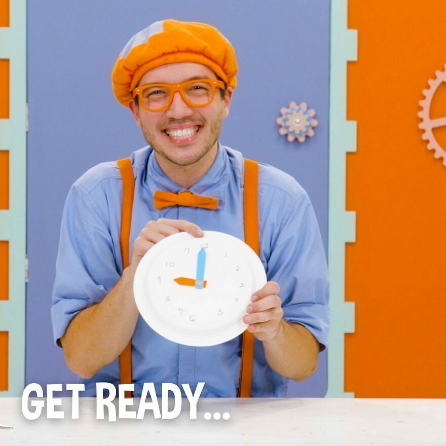 Smiling actor dressed as Blippi wearing orange bow tie, suspenders, and glasses, holding a paper clock in colorful setting.