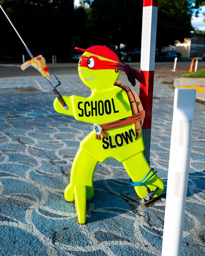 Bright yellow school slow sign dressed as a ninja turtle holding a paper cutout pizza slice in a street setting.